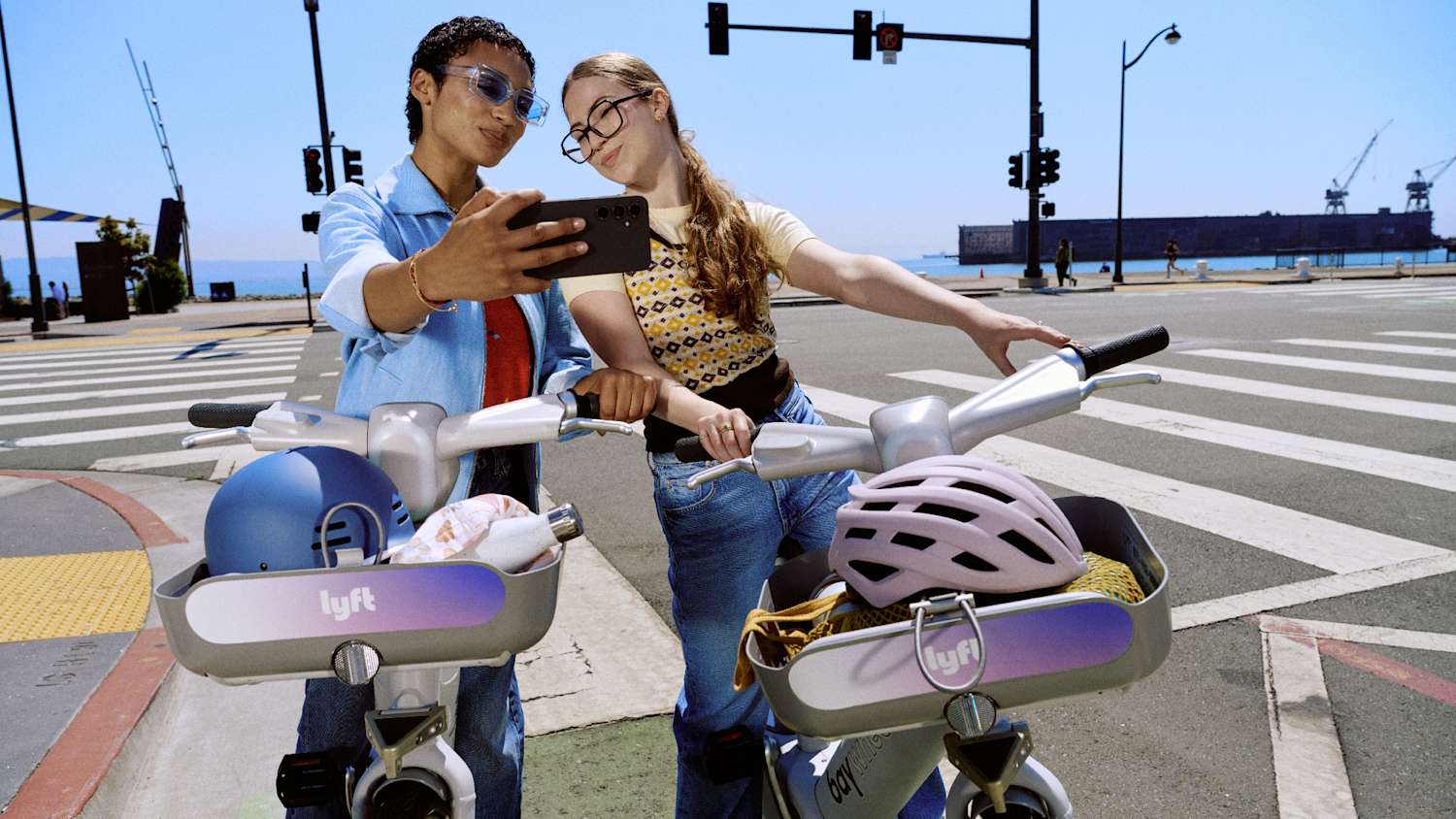 Two people take a selfie with Lyft bikes at a sunny city crosswalk, helmets in bike baskets.