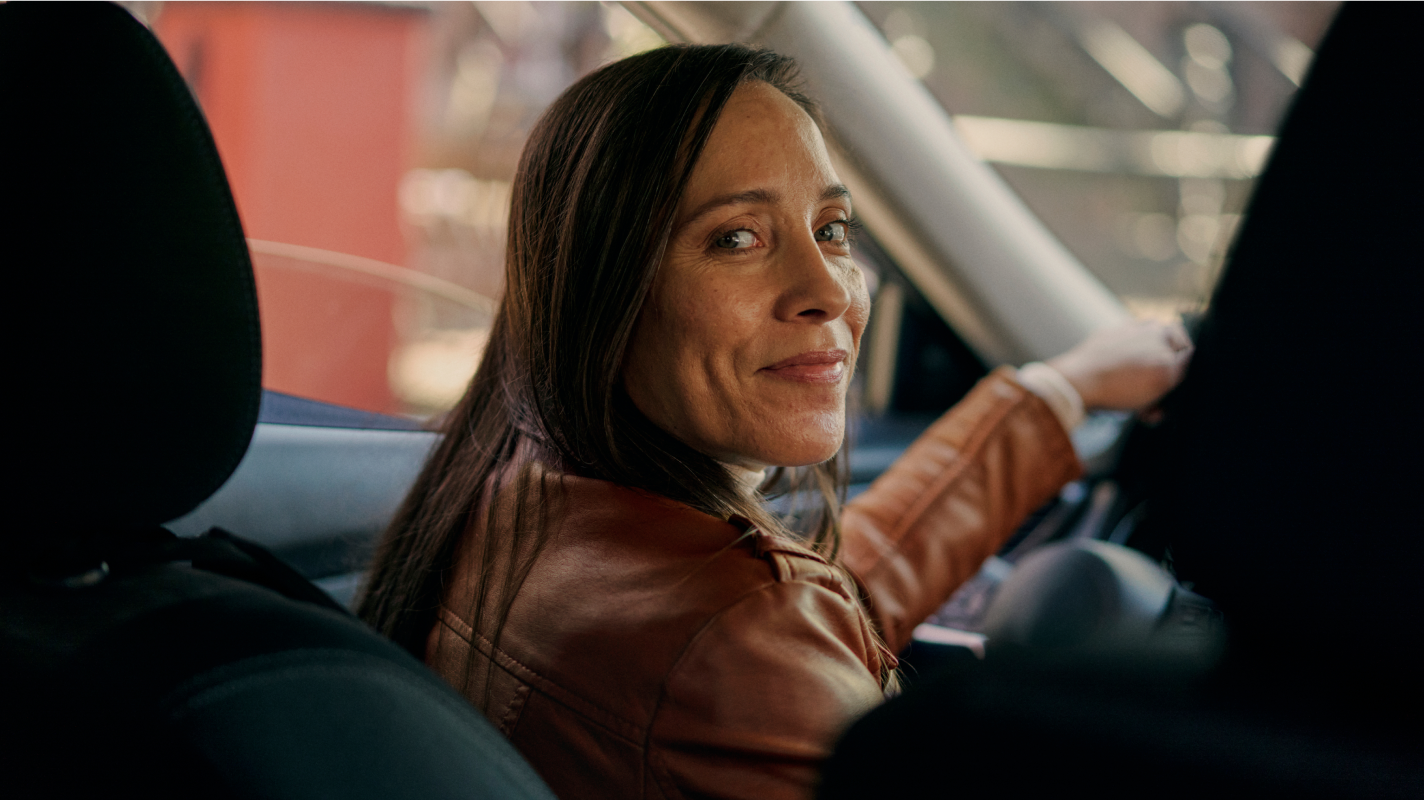 Woman in a brown leather jacket sitting in the driver's seat of a car, looking back over her shoulder and smiling. 