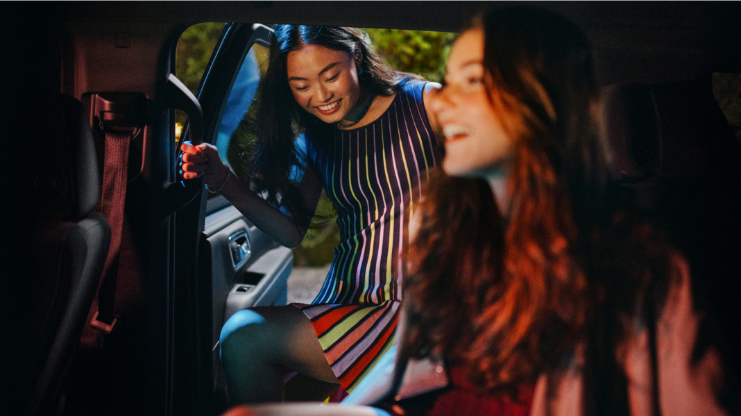 Two young women smiling, one entering a car wearing a colorful striped dress, with warm lighting inside the vehicle.