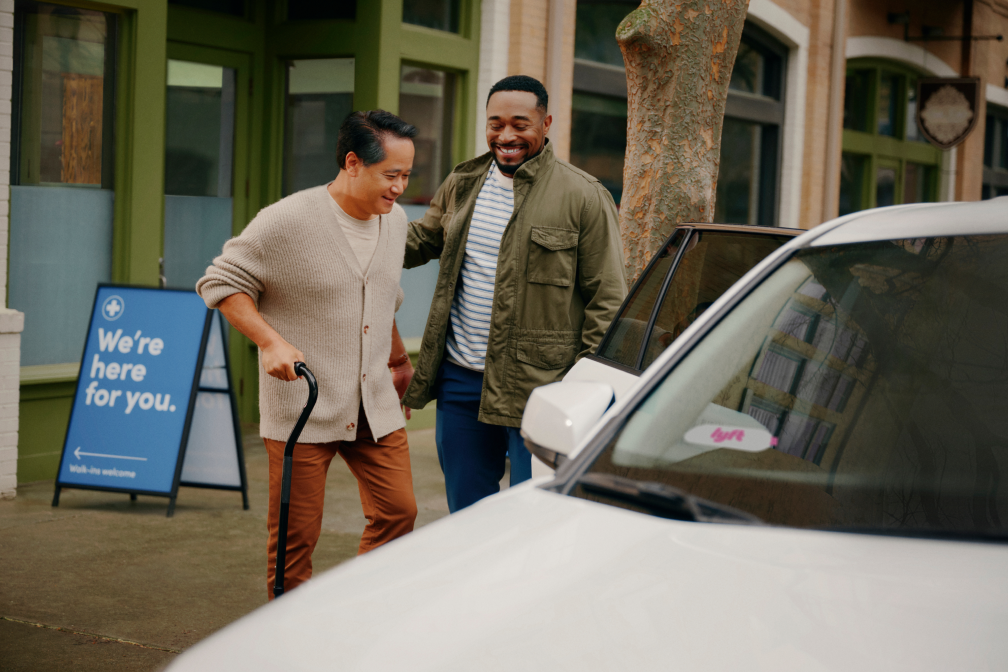 Two men smile as one helps the other, who uses a cane, get into a white car outside a building with a blue sign.