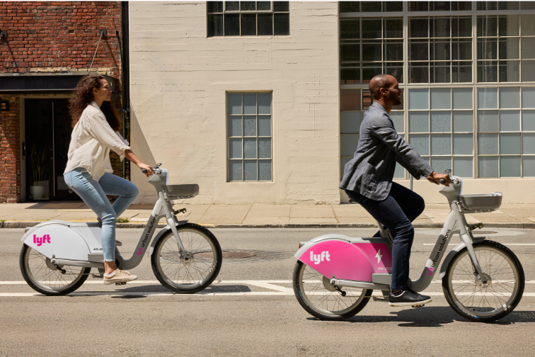 A woman and a man ride Lyft bikes on a city street in daylight, passing buildings with brick and cream-colored walls.