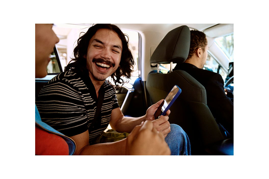Smiling man holding a phone in the backseat of a car, talking with a friend, driver in front.